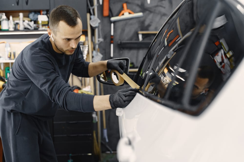 A man wearing a black shirt is fixing a car window, emphasizing vehicle care and the use of mirror stickers for protection.